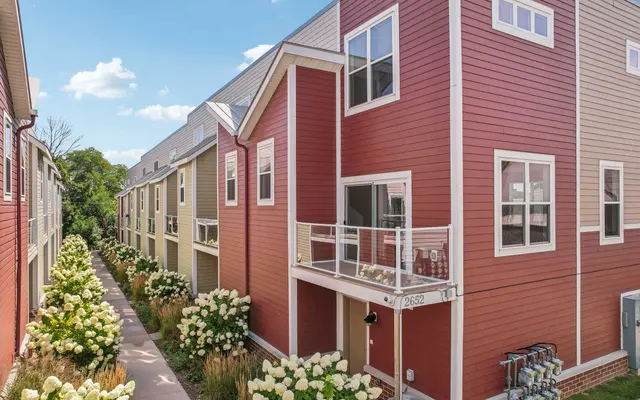 Exterior view of a modern townhouse complex with a pathway lined by flowering plants, featuring red and beige siding on the buildings.