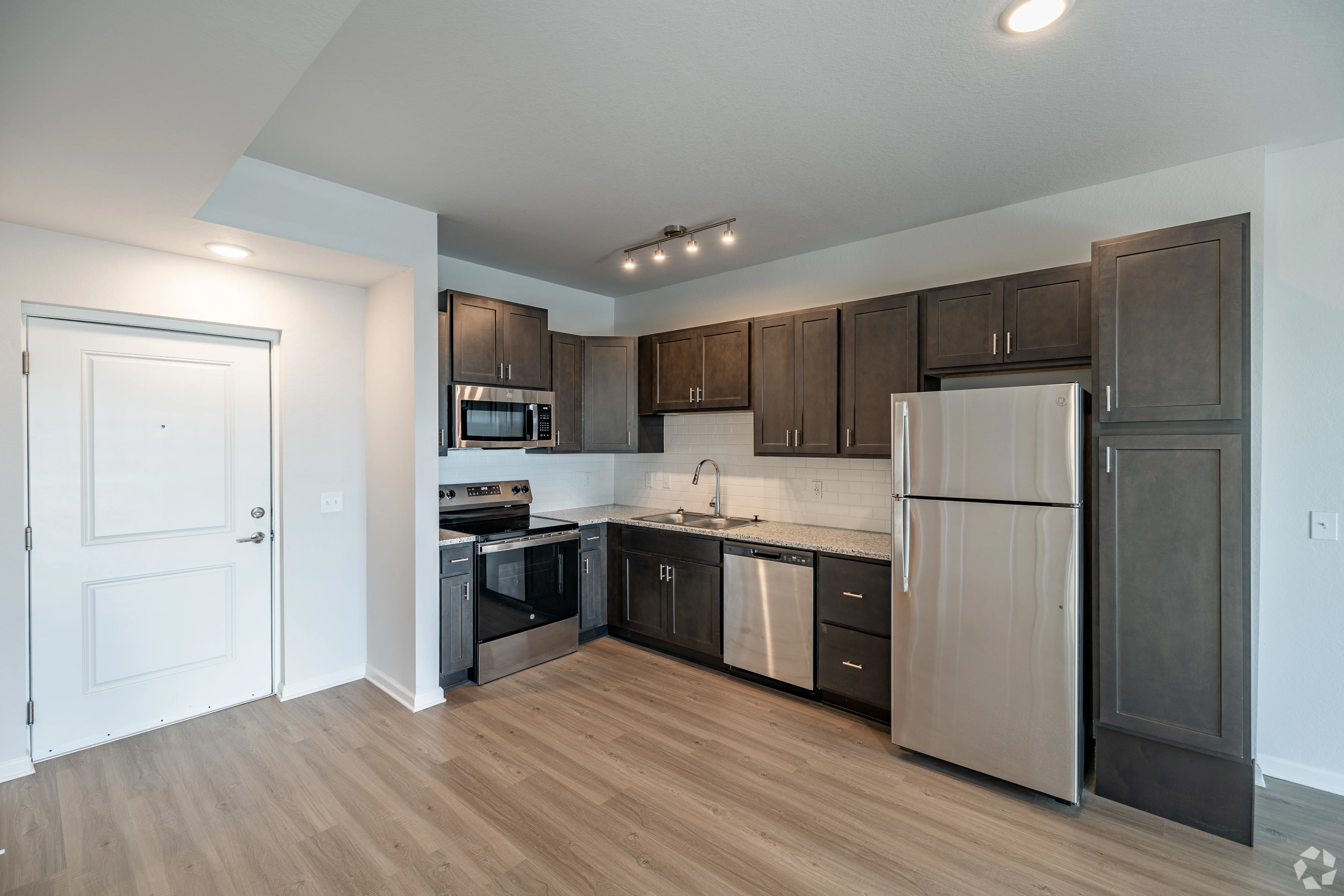 A modern kitchen featuring dark wood cabinets, stainless steel appliances, and light-colored countertops. The floor is made of light wood, and there is a door at the entrance.
