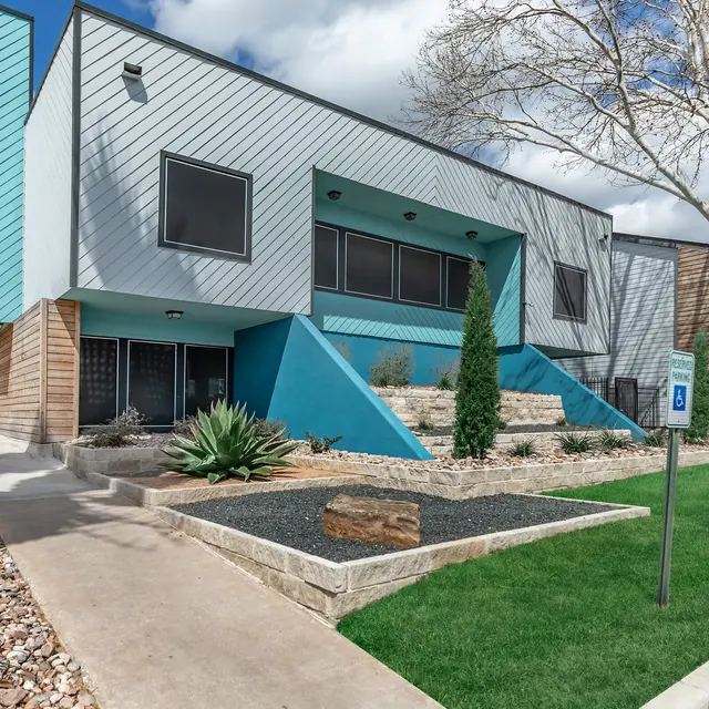 A modern building with a unique architectural design featuring a mix of blue and gray exterior walls. It has a landscaped area with plants and rocks in the foreground, along with a pathway leading up to the entrance.
