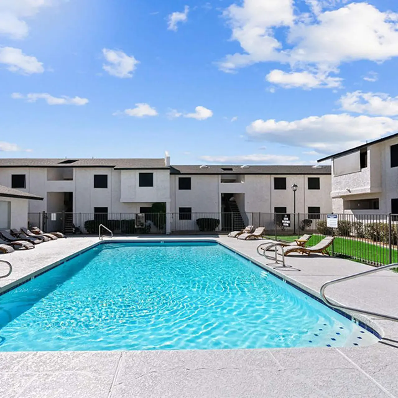 A swimming pool surrounded by lounge chairs and a fence, with apartment buildings in the background under a blue sky.