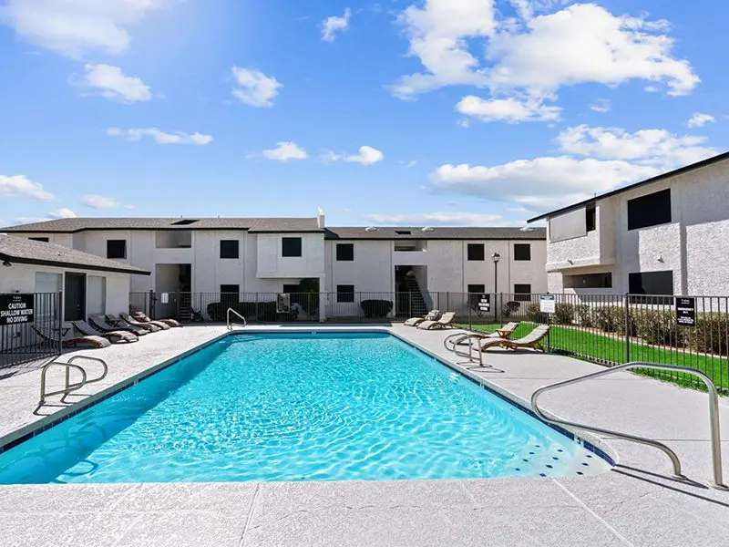 A swimming pool surrounded by lounge chairs and a fence, with apartment buildings in the background under a blue sky.