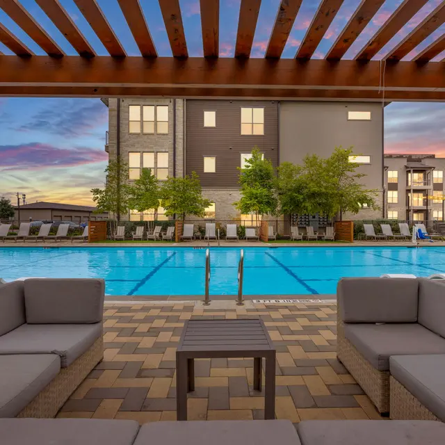 View of a swimming pool surrounded by lounge chairs and greenery, framed by a wooden pergola at sunset.