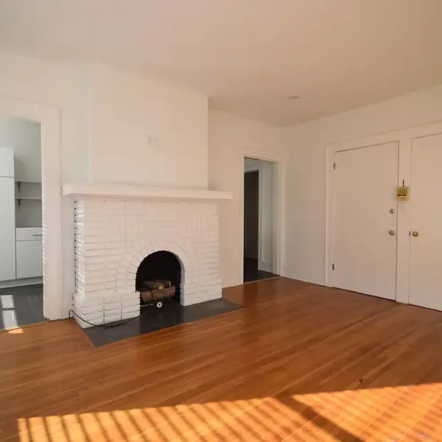 A spacious living room featuring a white fireplace and hardwood flooring. There are two doors leading to other rooms, and a glimpse of a kitchen area visible in the background. Natural light creates shadows on the floor.