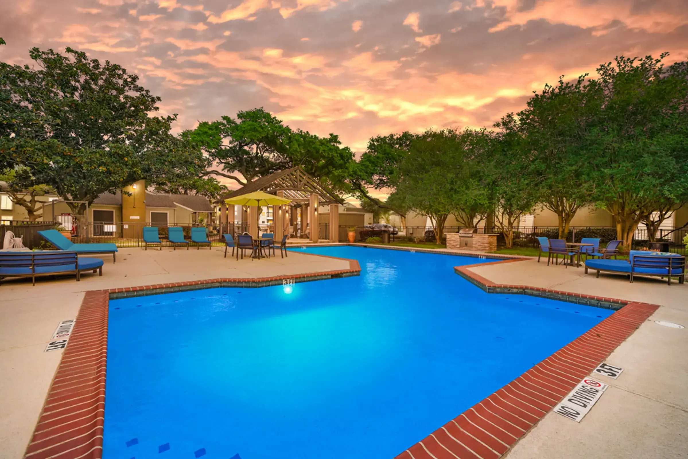 A beautifully illuminated swimming pool area at sunset, surrounded by trees and lounge chairs.