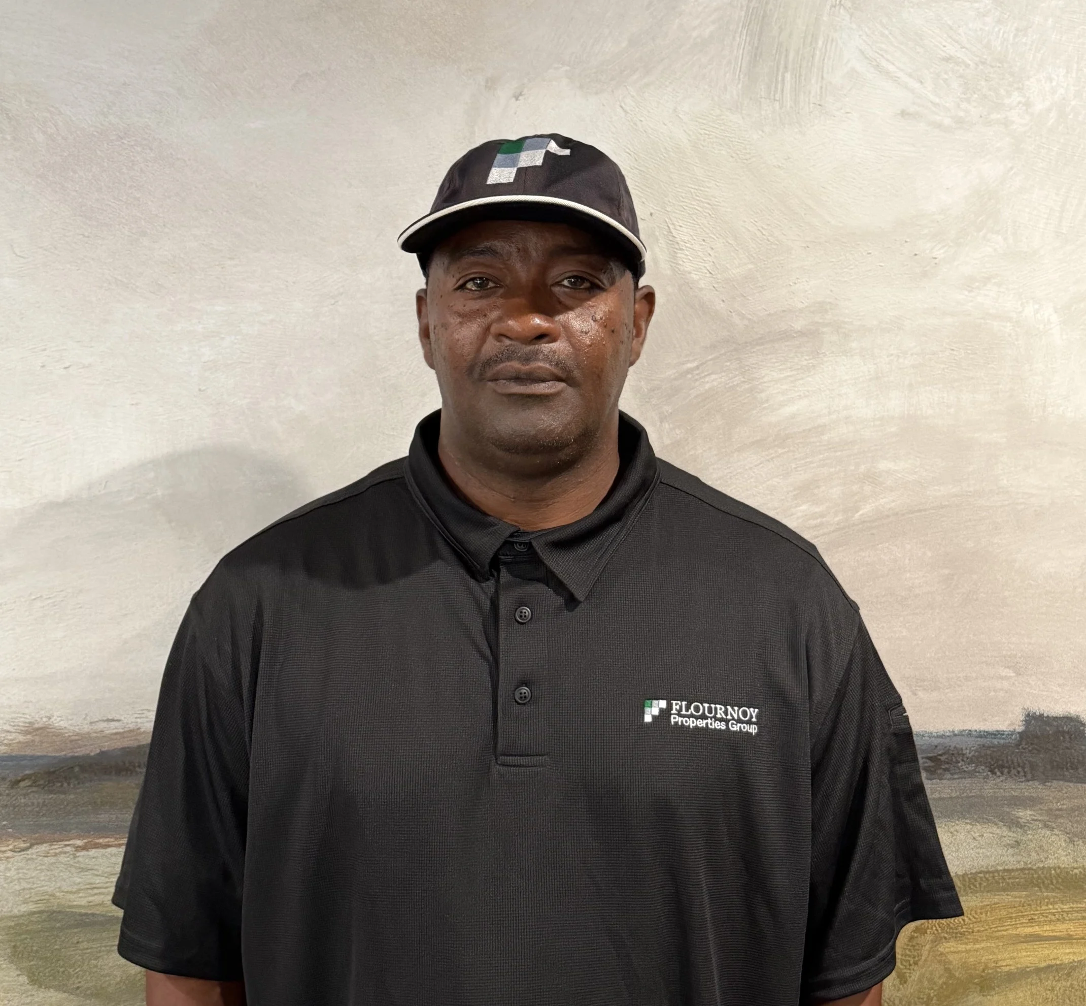 Staff Member Portrait A man standing in front of a mural wearing a black polo shirt and cap with a logo.