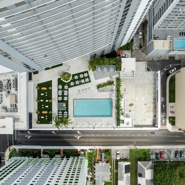 Aerial view of a modern building complex featuring a swimming pool and landscaped green areas, along with nearby roads and sports facilities.