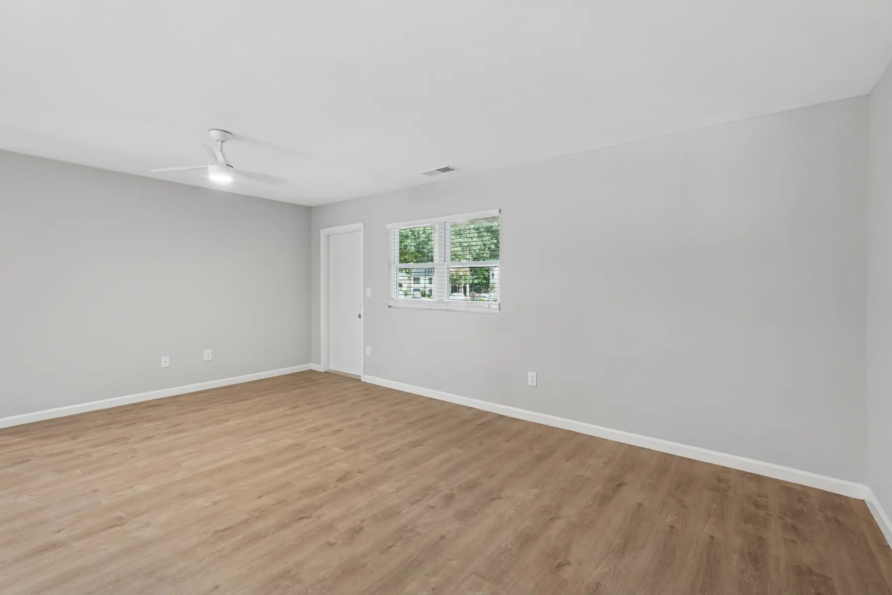 A spacious empty room featuring gray walls and light wood flooring. A ceiling fan is visible, and there are two windows allowing natural light to enter. A white door is on the left side of the room.