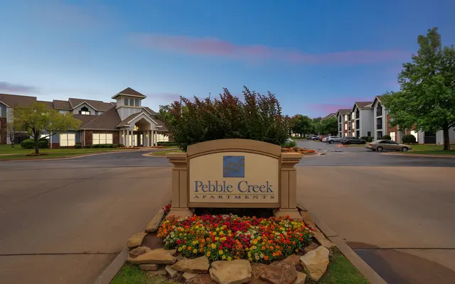 Sign for Pebble Creek Apartments surrounded by colorful flowers and greenery, with apartment buildings in the background under a twilight sky.