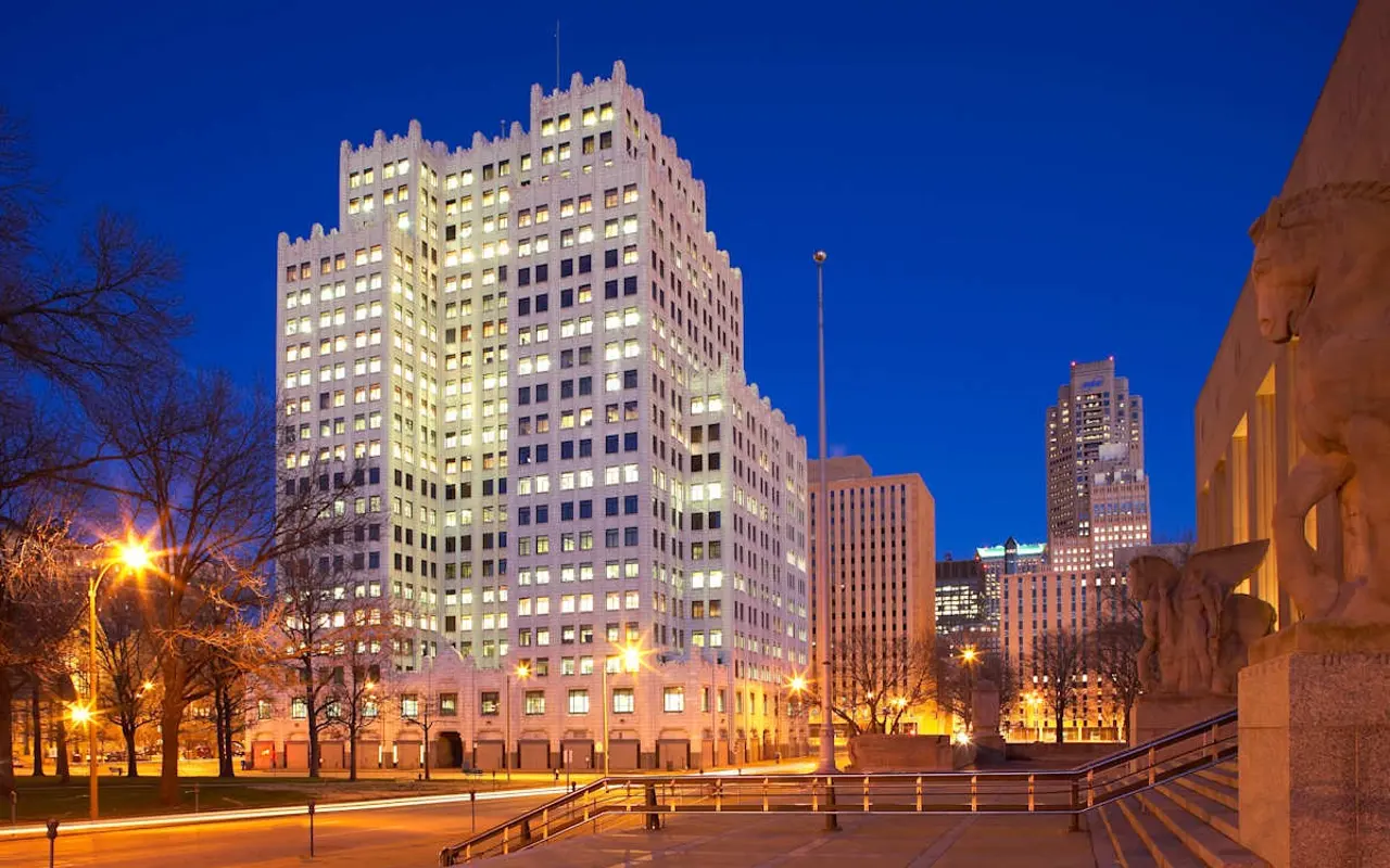 A lit-up skyscraper in an urban setting during the evening. The building features a modern architectural style with many windows reflecting lights. Surrounding trees are bare, indicating a season change. A stone staircase and statues can be seen in the foreground.