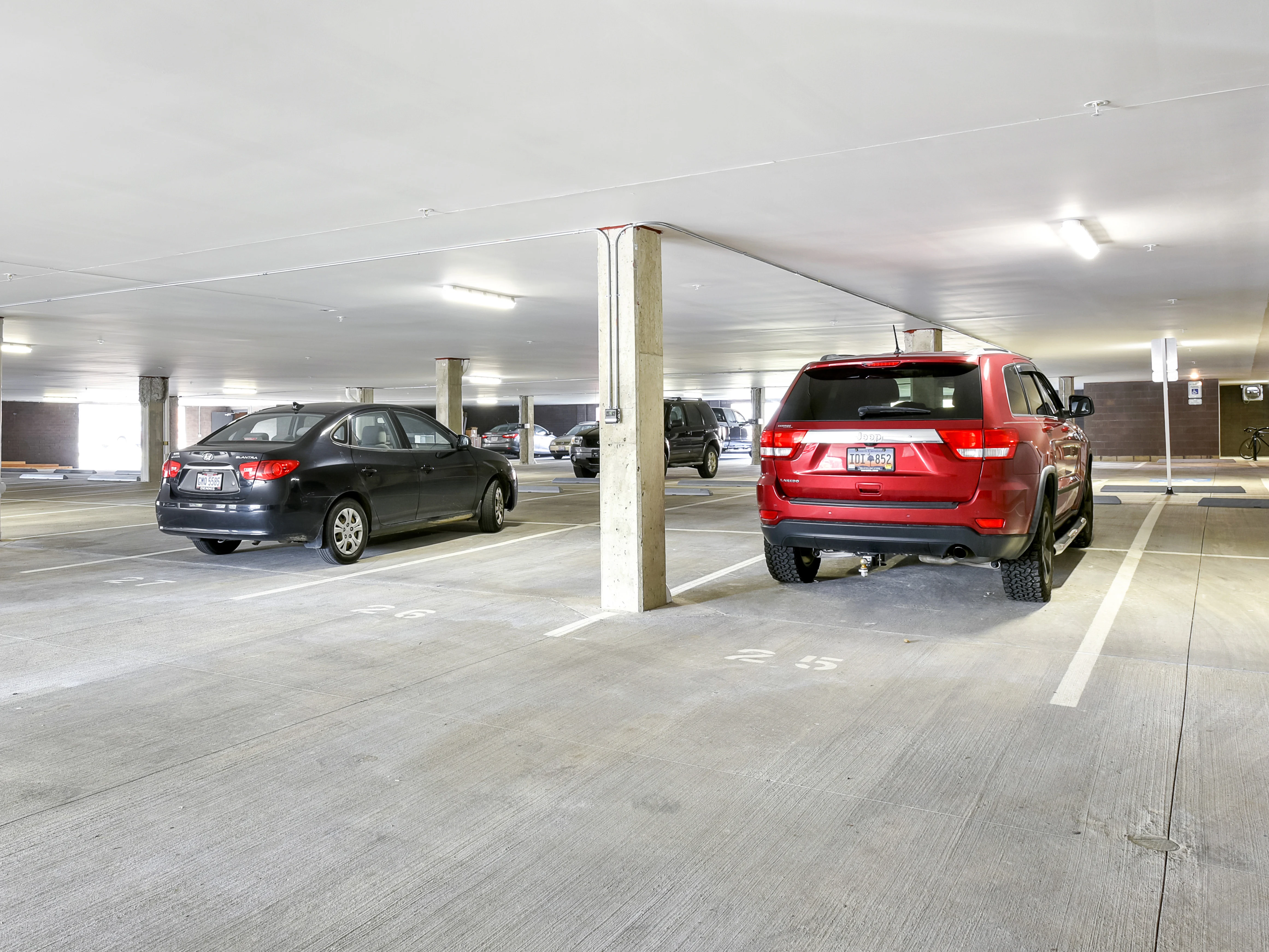 A spacious parking garage interior with several parked cars, including a red SUV and a black sedan. The ceiling is well-lit, and concrete pillars are visible.