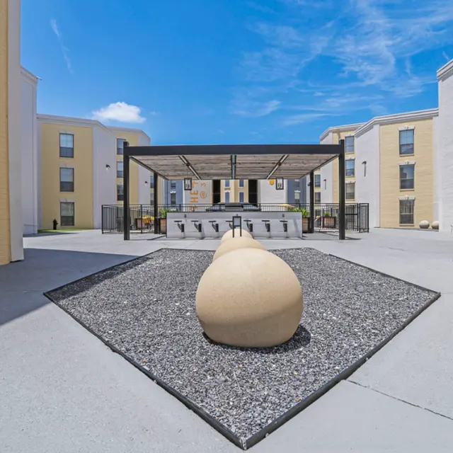 A spacious courtyard with a decorative stone feature in the center, surrounded by modern buildings. The area is sunny with a blue sky and some clouds visible. There is a shaded seating area with a roof structure in the background.