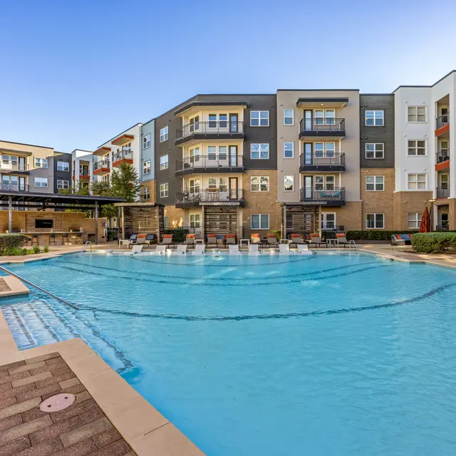 An outdoor swimming pool surrounded by apartment buildings, featuring lounge chairs and landscaped greenery.