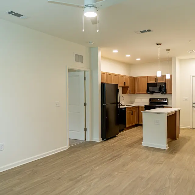 A modern kitchen and living area featuring wood cabinets, a black refrigerator, and an island with pendant lighting.