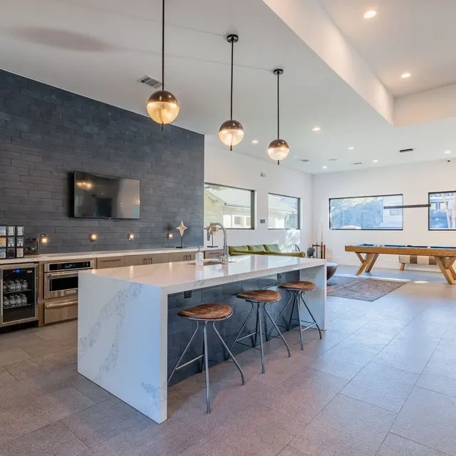A modern kitchen with an island, bar stools, and a dark accent wall. There is a television mounted on the wall, a dining table in the background, and large windows letting in natural light.