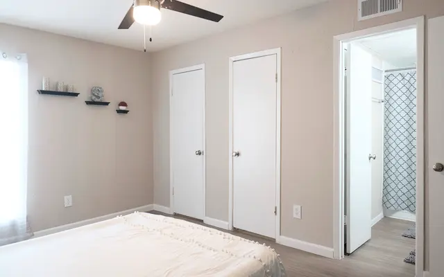 A minimalist bedroom with a bed in the foreground, light beige walls, and two closed white doors. A small shelf with decor items is mounted on the wall, and there's a doorway leading to a bathroom visible in the background.