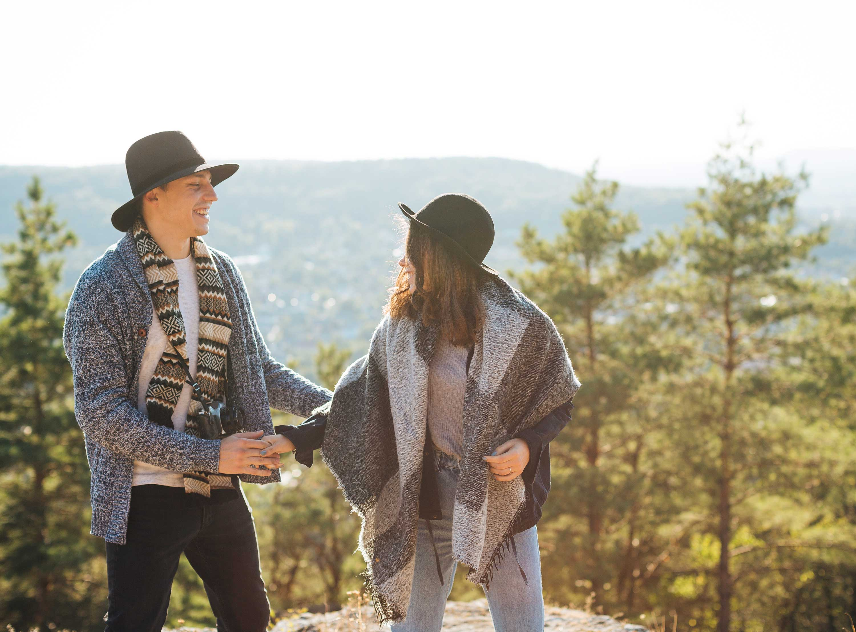A couple standing outdoors on a sunny day, wearing hats and cozy scarves. They are smiling and holding hands, surrounded by trees and a distant view of hills.