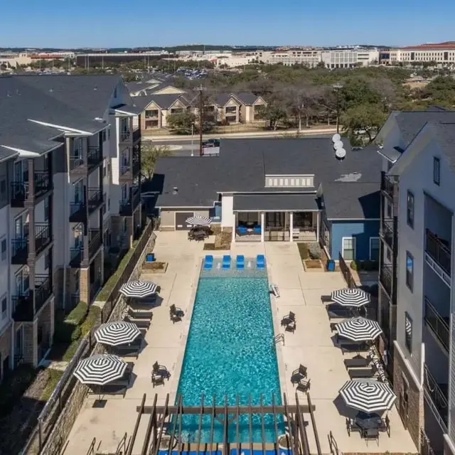 Aerial view of a modern apartment complex featuring a large swimming pool surrounded by lounge chairs and umbrellas.