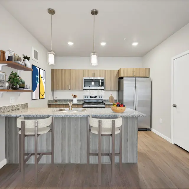Modern Kitchen Interior A modern kitchen featuring a central island with bar seating, equipped with stainless steel appliances and wooden cabinets. The space is well-lit with pendant lights and decorated with a small plant and a bowl of fruit on the island.