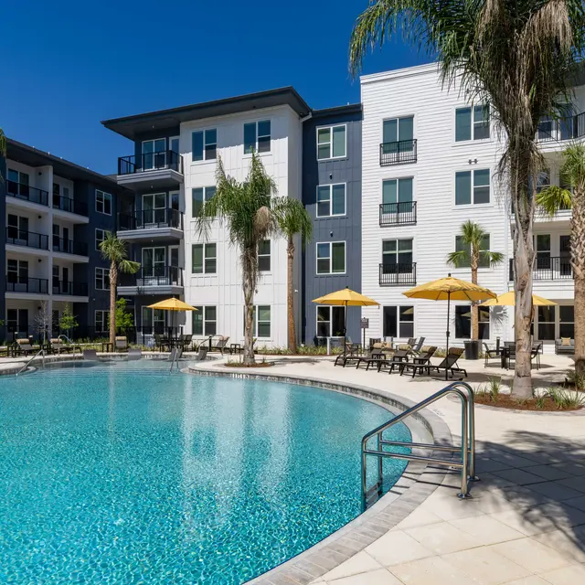 A modern apartment complex featuring a swimming pool, surrounded by palm trees and lounge chairs with umbrellas.