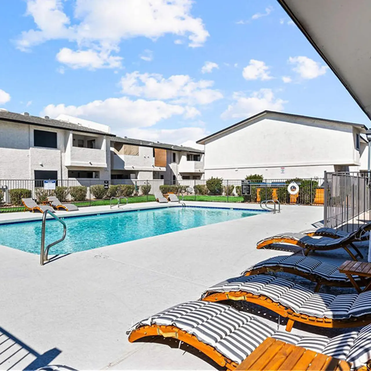 A sunny pool area surrounded by apartments, featuring lounge chairs and a clear blue sky.