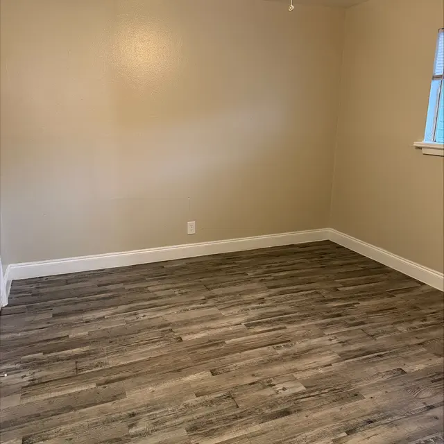 A spacious empty room with light-colored walls and laminate flooring. A ceiling fan is visible, and there's a window allowing natural light to enter the space.