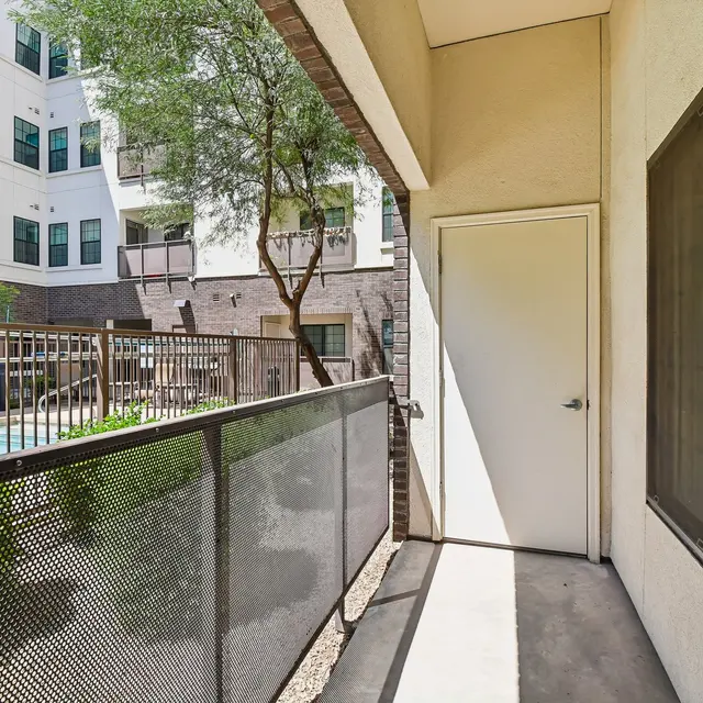 Apartment Balcony View A balcony view of an apartment complex with a pool in the background. The balcony features a mesh railing and a door leading inside. Surrounding the pool are various trees and the exterior of the building with large windows.