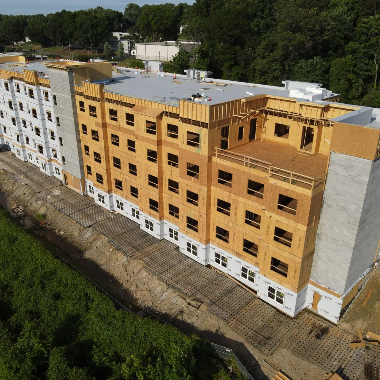A multi-story building under construction, featuring wooden framing and partially finished exterior. Surrounding area includes dirt and greenery.