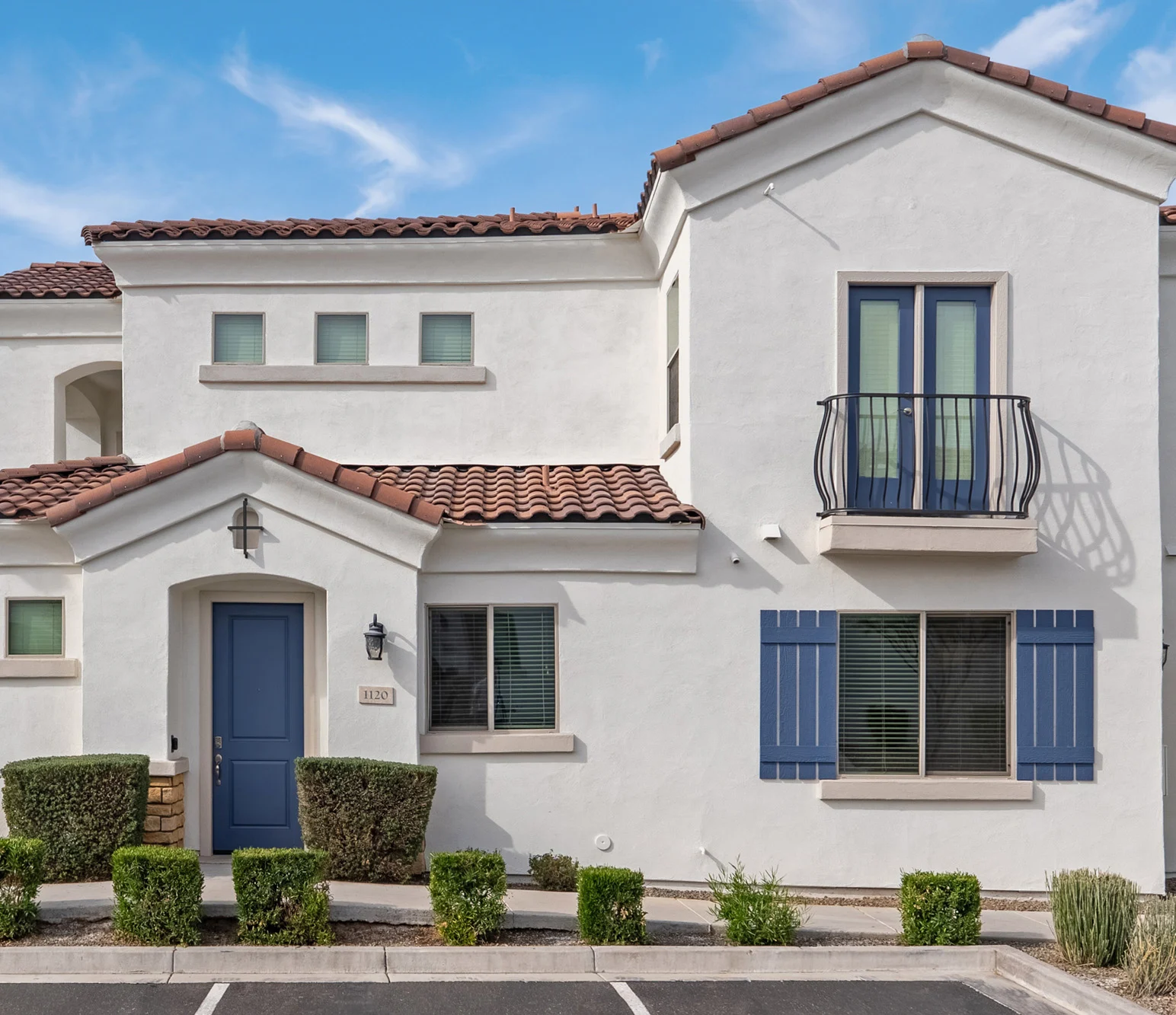 A modern two-story house with white stucco walls, a red-tiled roof, and blue shutters. The front features a blue door and a small balcony on the upper floor.