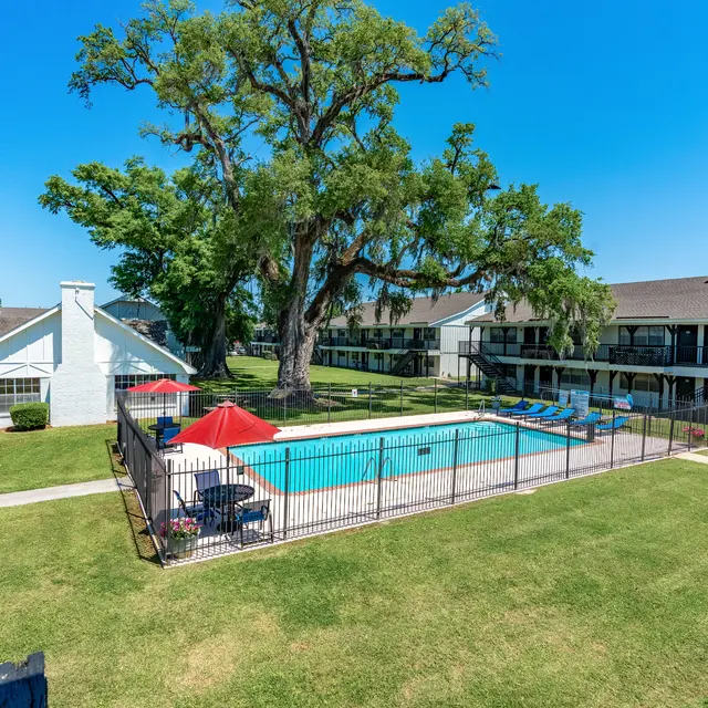 Aerial view of an apartment complex featuring a swimming pool surrounded by grass and lounge chairs, with parking spaces visible in the background and large trees providing shade.