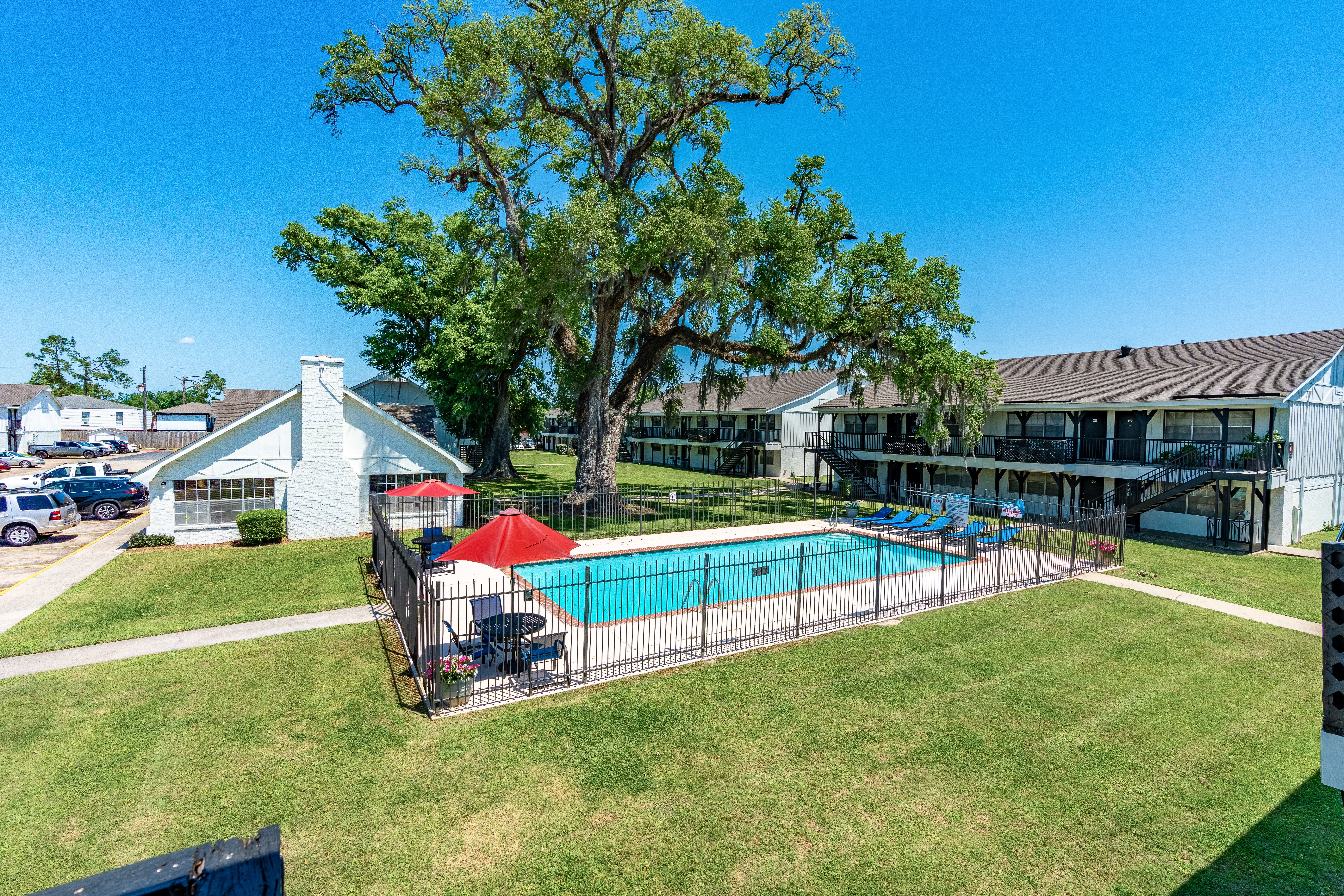 Aerial view of an apartment complex featuring a swimming pool surrounded by grass and lounge chairs, with parking spaces visible in the background and large trees providing shade.