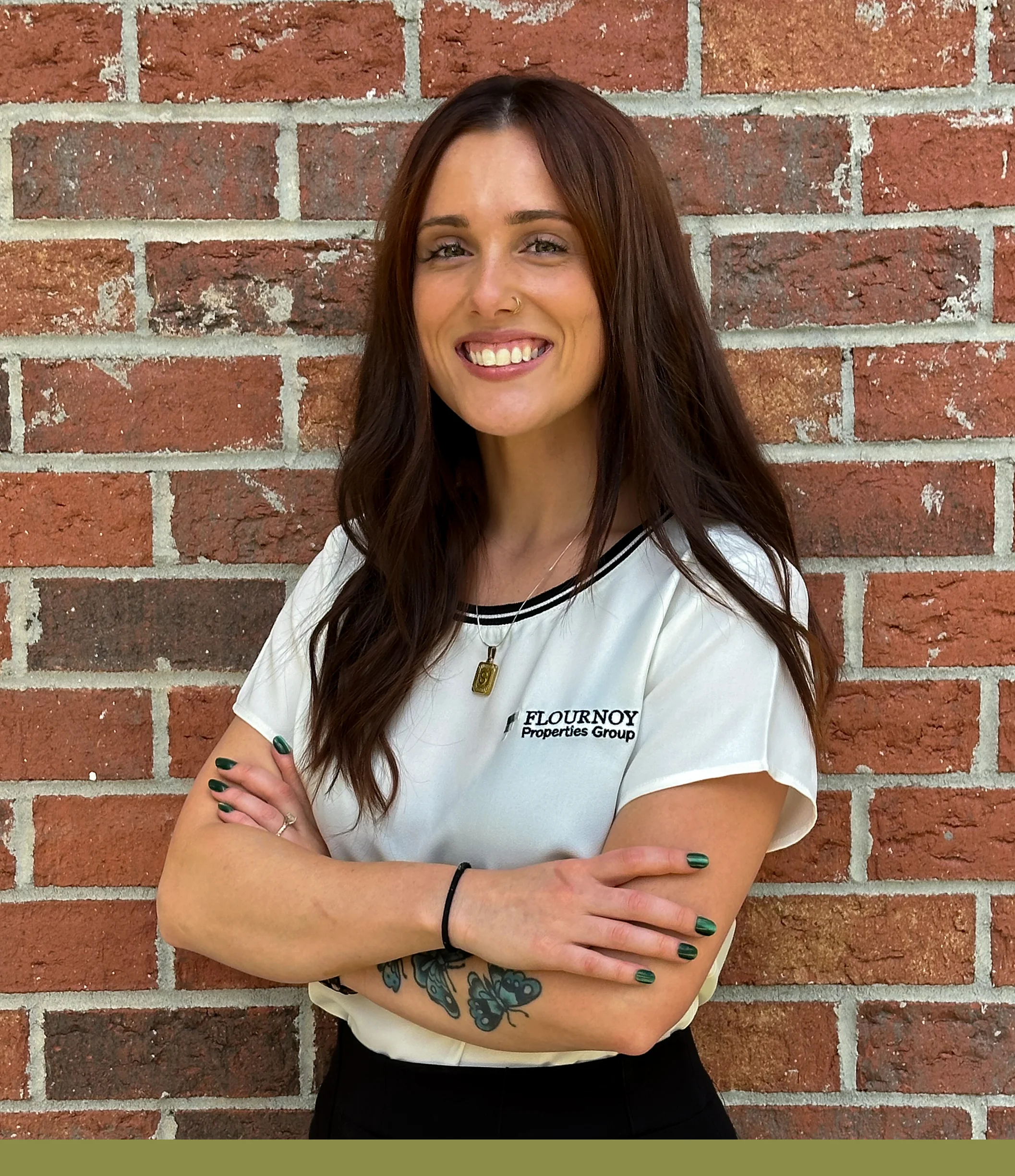 Professional Portrait of a Woman A woman smiling while standing against a brick wall. She has long brown hair, wearing a white shirt with a black collar and black pants, and has her arms crossed confidently.