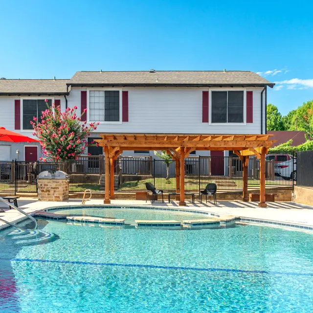 A vibrant swimming pool area featuring a wooden pergola and lounge chairs near the pool. Behind it, there are two-story residential buildings with a red umbrella and greenery surrounding the area.