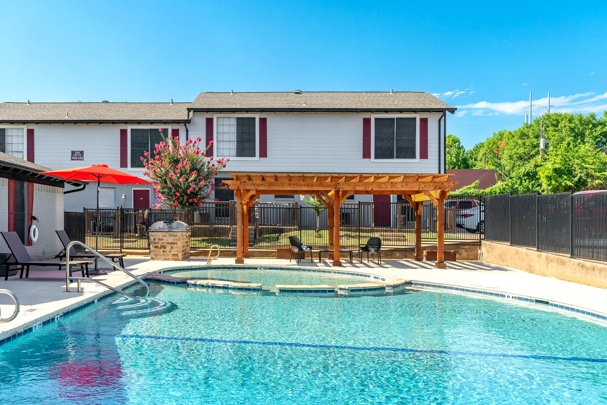 Swimming Pool Area with Pergola A vibrant swimming pool area featuring a wooden pergola and lounge chairs near the pool. Behind it, there are two-story residential buildings with a red umbrella and greenery surrounding the area.