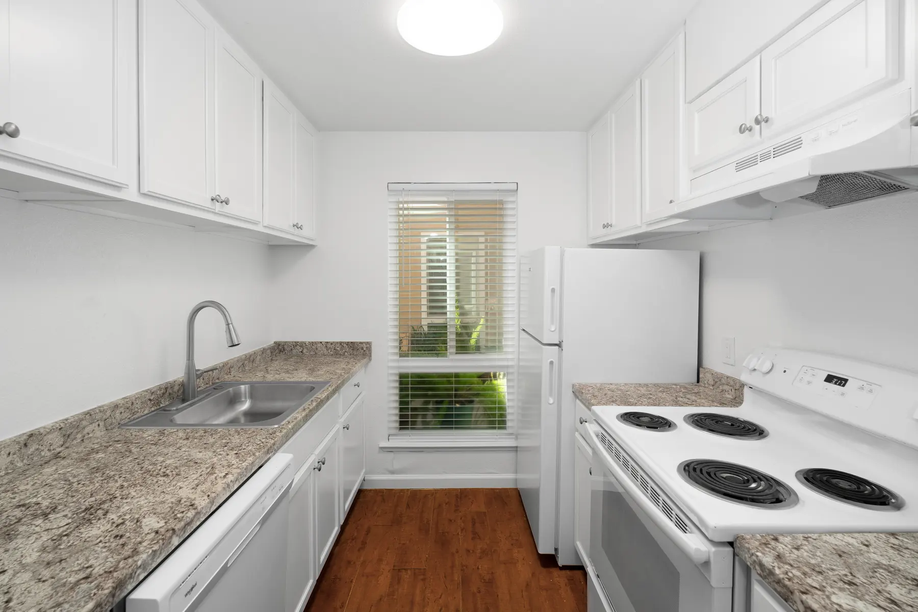 A modern kitchen featuring white cabinetry and granite countertops, with a stainless steel sink and a white stove and fridge. The room is well-lit and includes a window with blinds.