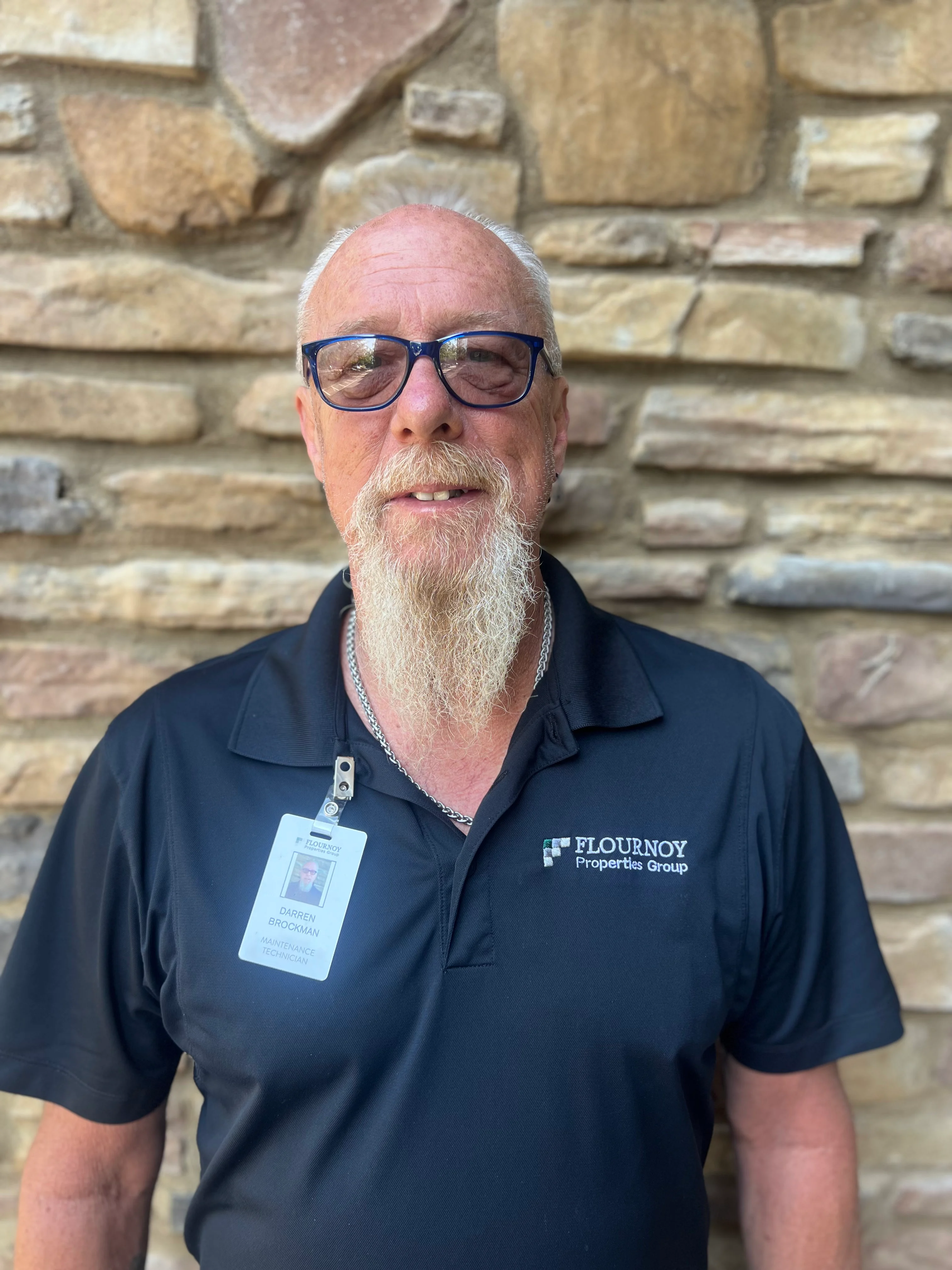 A man with a white beard and glasses, wearing a black polo shirt with a logo, standing against a stone wall.