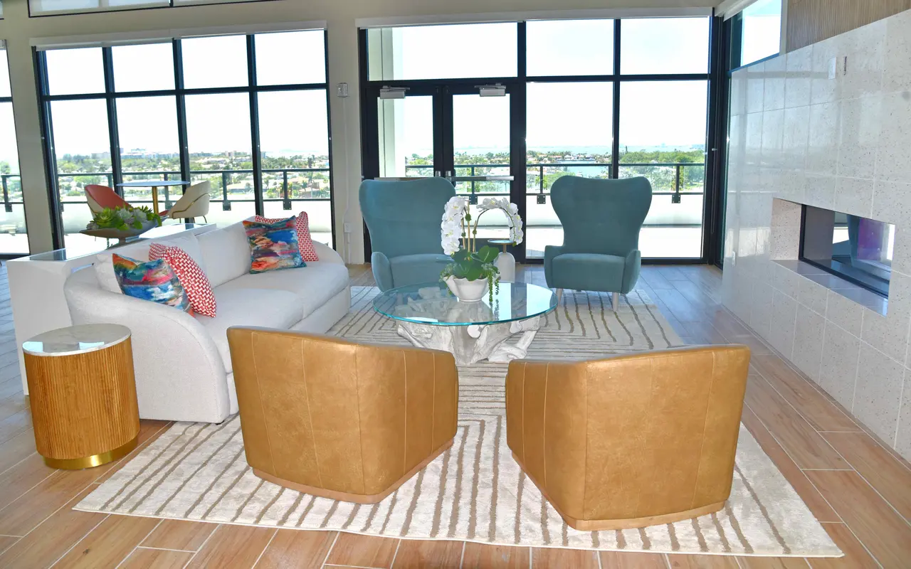 A modern living room featuring a white sofa, two blue chairs, and two tan armchairs arranged around a glass coffee table on a striped rug. Large windows provide a view of the outside.