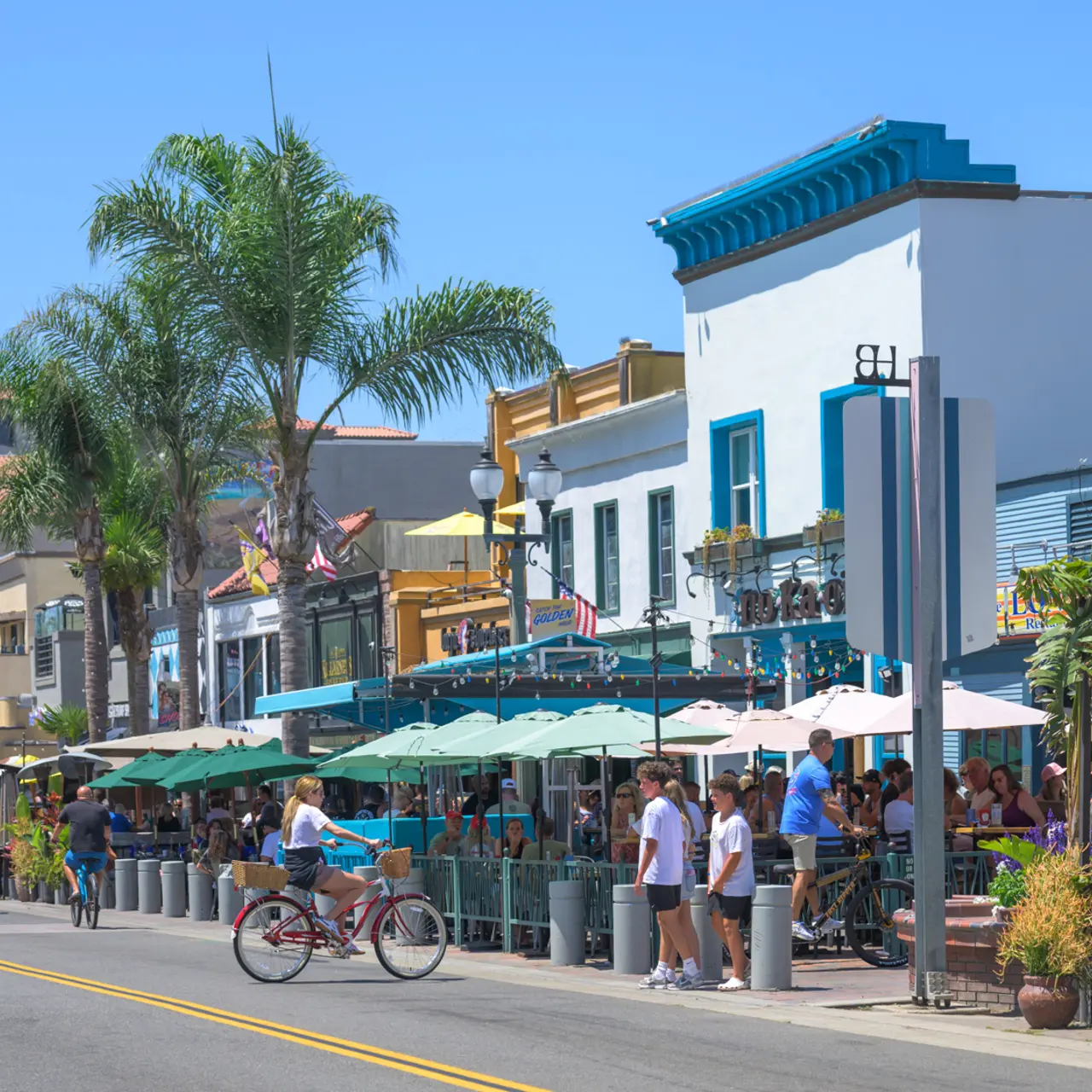 A vibrant street scene featuring outdoor dining areas with umbrellas, palm trees, and people biking along the sidewalk on a sunny day.