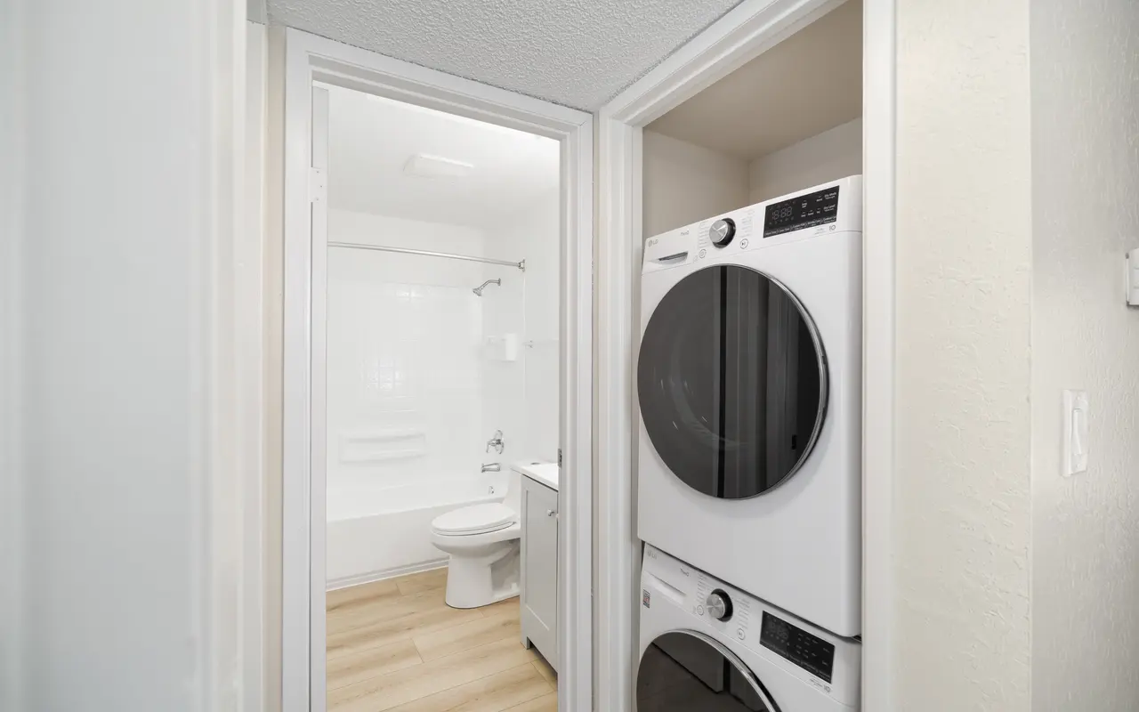 A view of a laundry room and adjacent bathroom, featuring stacked white washing machines and a bathroom with a shower and toilet in background.