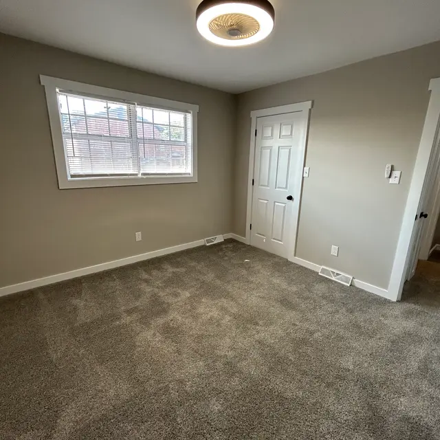 A spacious empty room with light gray walls, a carpeted floor, and a large window allowing natural light in. The ceiling features a modern light fixture. There is a closed white door on one wall.