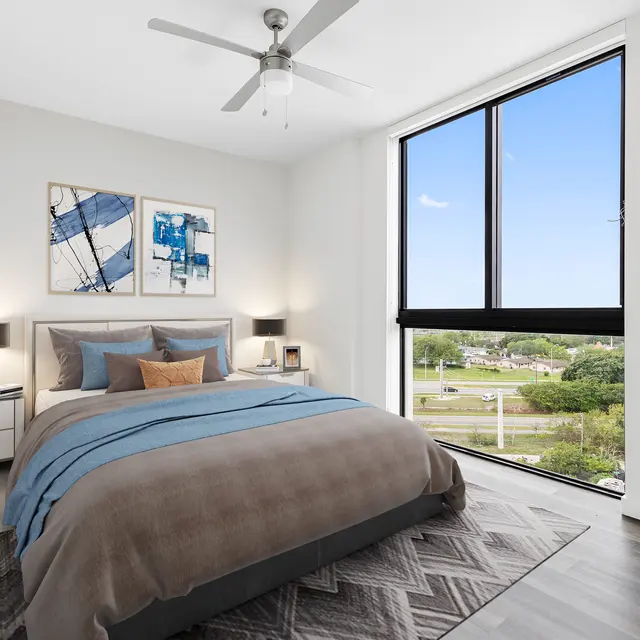 A modern bedroom with a large window showing an outdoor view. The room features a comfortable bed with gray and blue bedding, a plant, and wall art.
