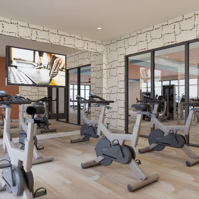 Interior of a modern gym with stationary bikes, large windows, and a TV on the wall showing a workout instruction video.