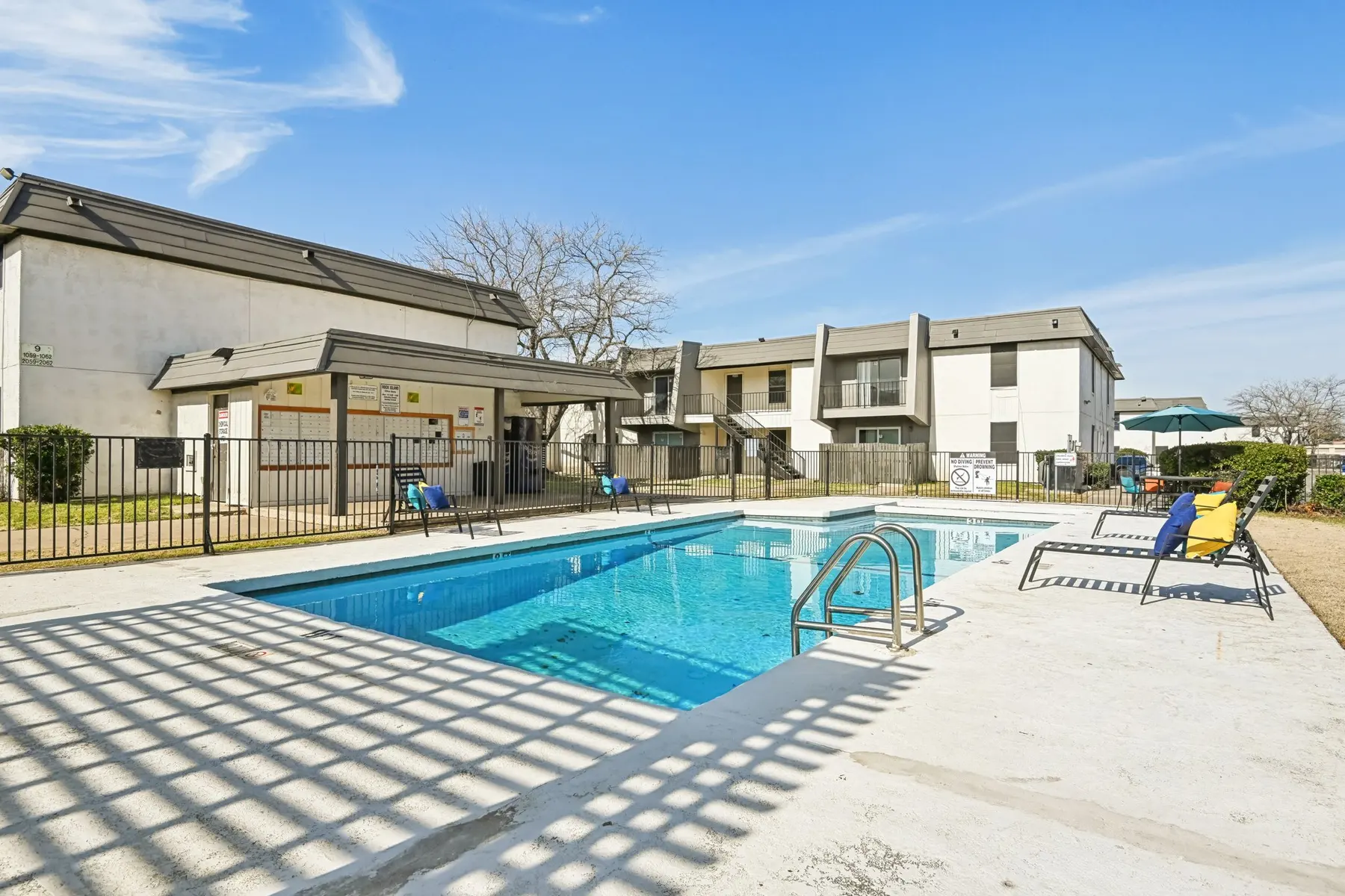 A swimming pool area in an apartment complex with lounge chairs and a shaded cabana. The pool is clear and surrounded by a concrete deck and safety fence, with apartment buildings in the background.