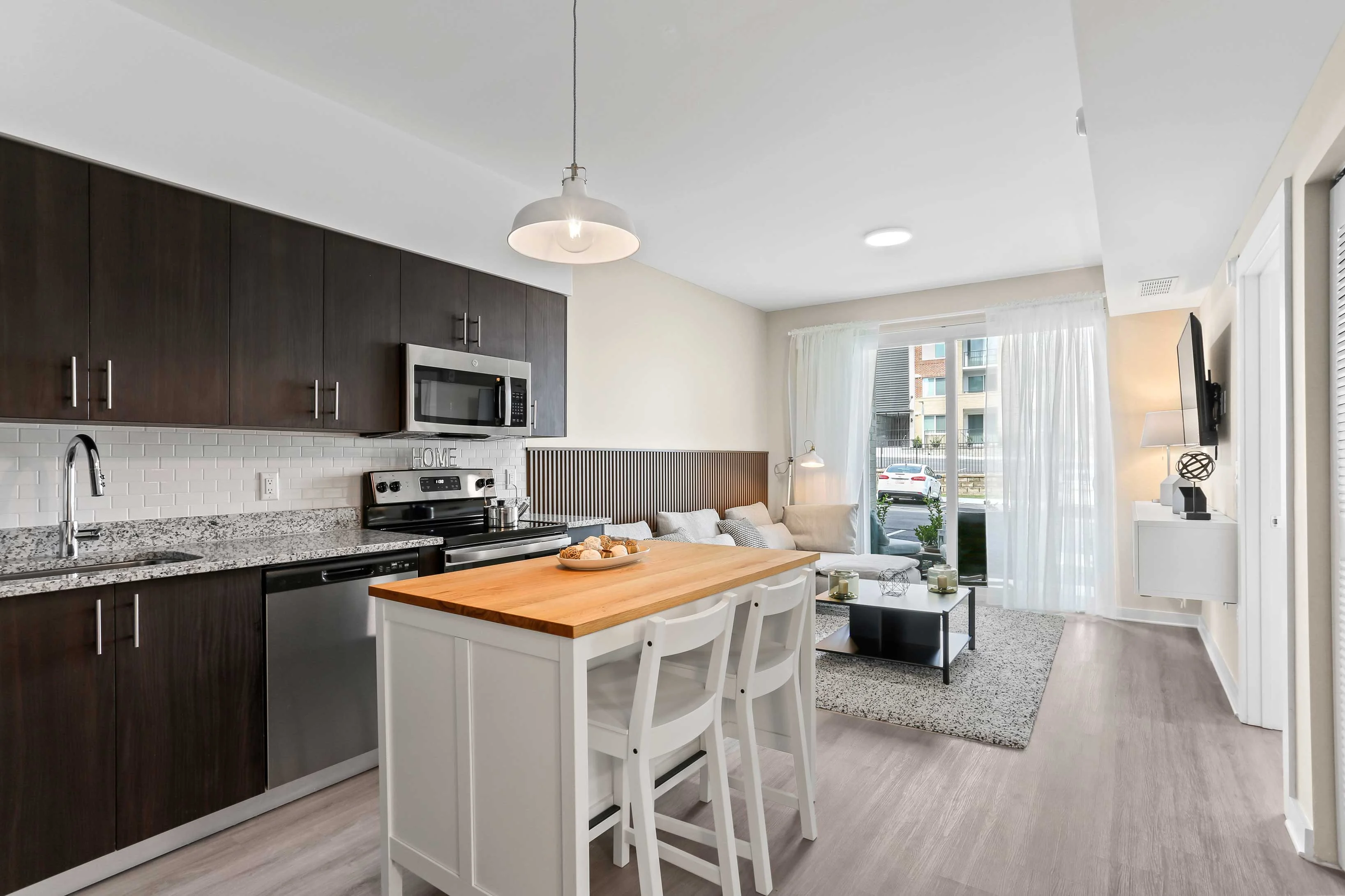 Modern Apartment Kitchen and Living Room A modern kitchen and living room area featuring dark cabinetry, stainless steel appliances, and a wooden dining table with white chairs. Natural light streams in from large sliding glass doors leading to a patio. The decor is stylish and minimalist, with a cozy seating area visible.
