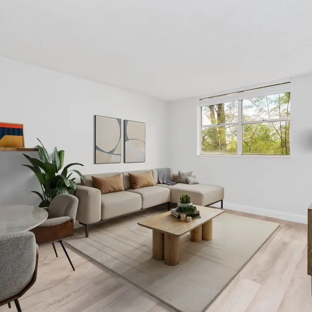 A modern living room featuring a light-colored sofa, a round dining table with upholstered chairs, and a wooden coffee table. The walls are white, adorned with abstract art. A window offers a view of greenery outside.