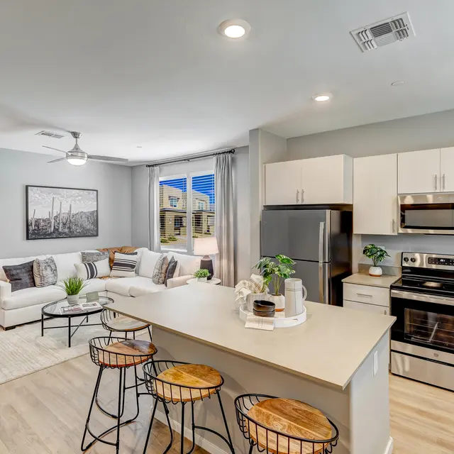 Modern living room and kitchen with a white sofa, dining area, stainless steel appliances, and bright natural light.