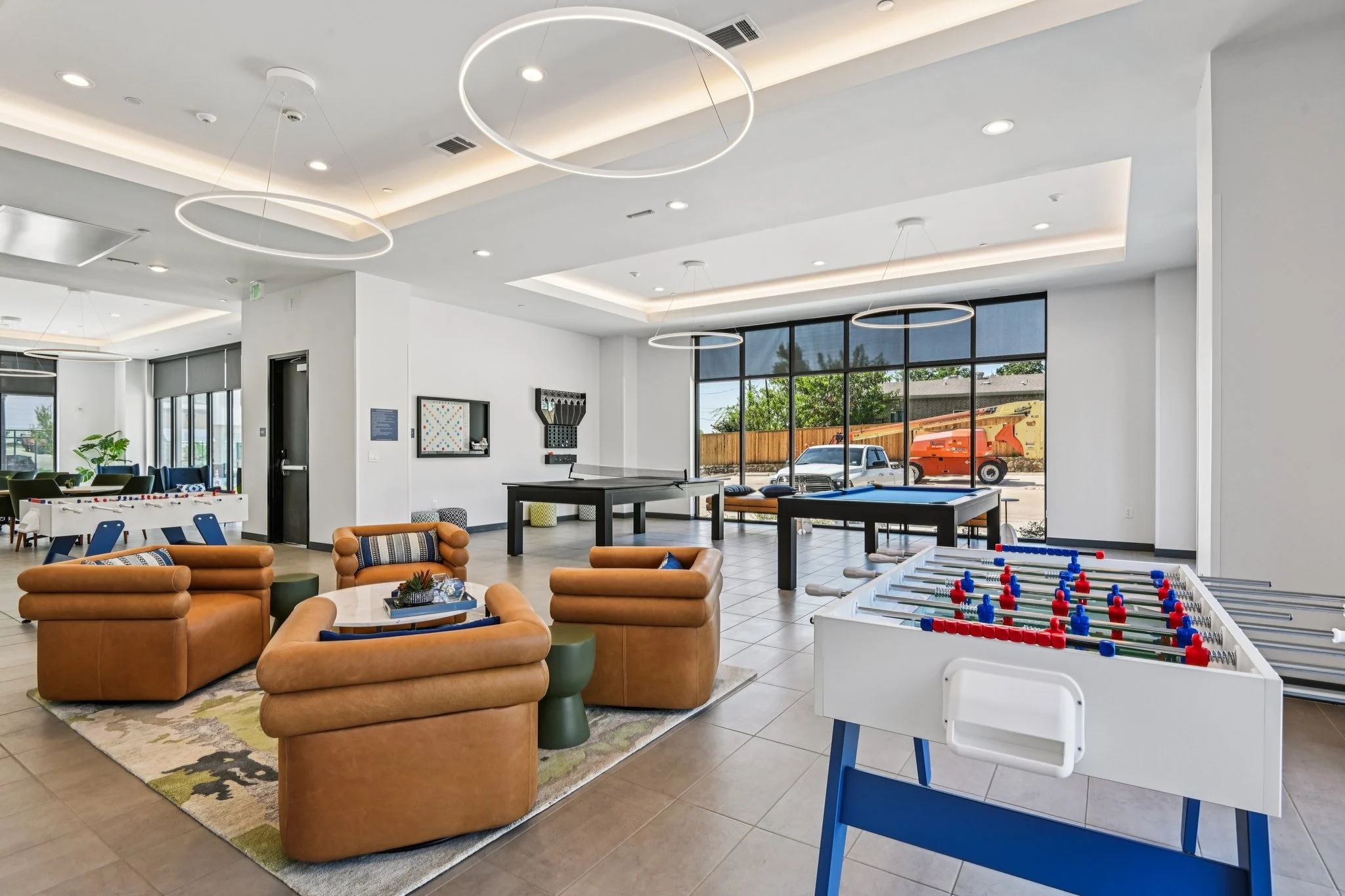 A spacious community lounge area featuring seating, a foosball table, and large windows with a view outside.