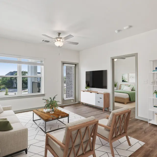 A modern living room featuring a comfortable sofa, two armchairs, a wooden coffee table, and a TV console. Large windows provide ample natural light, and a white shelving unit adds a touch of decor. A door leads to a bedroom in the background.