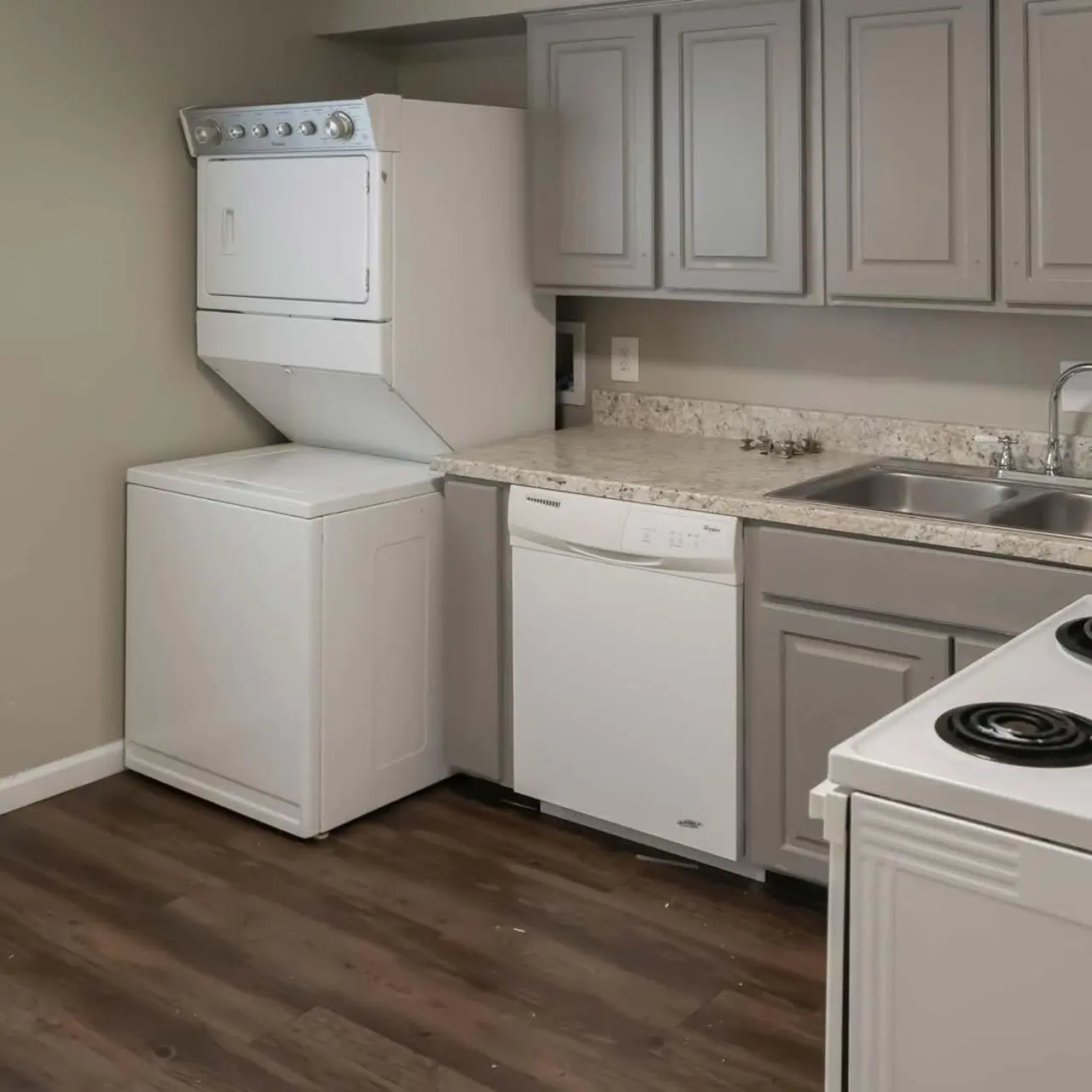 A modern kitchen featuring a stacked washer and dryer, a white dishwasher, and a double sink with a faucet. The kitchen has light gray cabinets and a stove with four burners. The floor is made of brown hardwood-like material. Natural light enters from a window on the left.