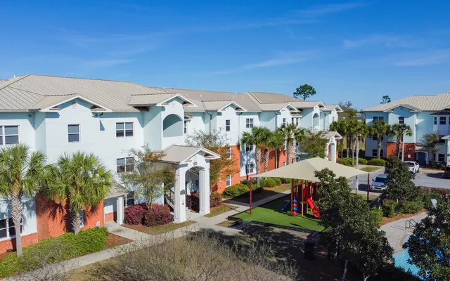 Apartment Complex with Playground Aerial view of a modern apartment complex surrounded by palm trees and greenery, with a playground area visible in the foreground.