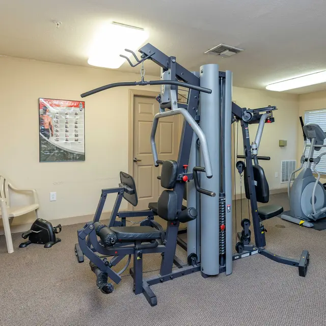 A small gym area featuring exercise equipment including a multi-function machine, an elliptical trainer, and a stability ball. A chair and some personal items are visible in the background.